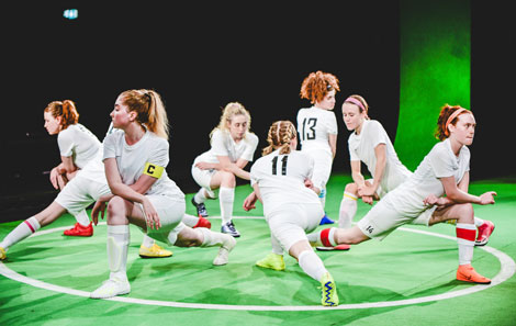 A scene from The Wolves, designed by Rose, showing the female football team in white kit, in a circle facing outwards, stretching their thigh muscles 
