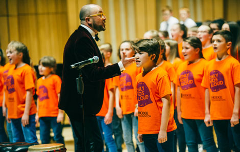 Tim, in a tuxedo conducts a group of Only Kids Aloud choir in bright orange t shirts 