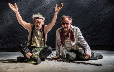 Ian McKellen as King Lear in a production directed by Jonathan Munby. He's dressed in rags with glassed on and has his hands in the air in an open gesture. Next to him is Gloucester, also in rags, with his eyes seemingly taken out. 