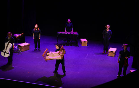 Flying Bedroom rehearsal - the students stand around the stage, one at the front with a model of a bed with a puppet in it, one with a cello, and the others singing 