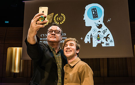 RWCMD Fellow Russell T Davies and Callum take a selfie in front of the screen advertising Russell's BAFTACymru talk in 2019