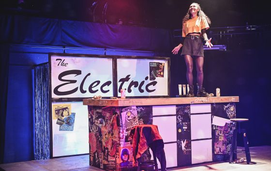 The Electric set: a girl stands on a bar and behind her is a wall with The Electric written on it 