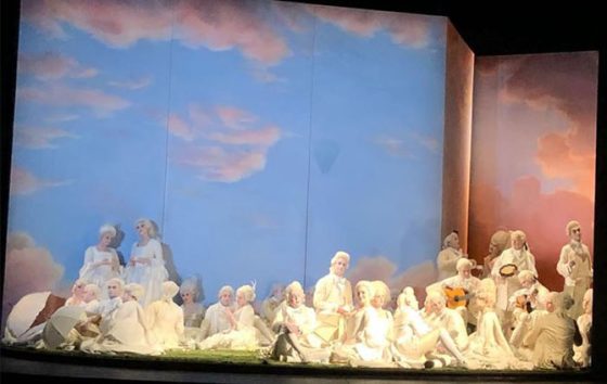 George on stage performing with members of Glyndebourne opera company - a backdrop of blue sky with a stage full of male and females dressed in white regency costume 