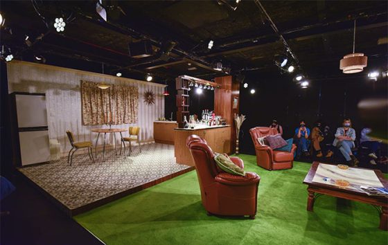 The set of Abigail's Party, with the lounge in the back half of the stage, and a green carpert with two comfortable chairs in the foreground. 