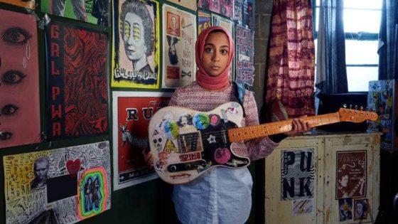 A still from the Channel 4 comedy We are Lady Parts. .Anjana is standing in front of posters on the wall, holding a guitar covered in graffitti 