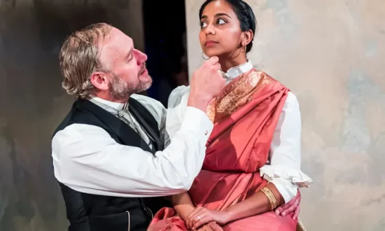 In a production photo from A Doll's House, Anjanaas Niru sits demurely on the knee of a man in old fashioned clothes as he chucks her under the chin 