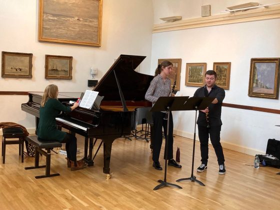 In a room lined with art work, Julien Bliss talks to a music student who has just performed, standing next to a Steinway grand piano 