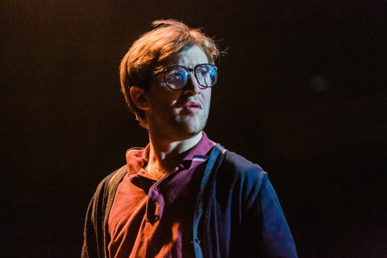 Callum in character, performing in RWCMD's production of Motortown. He's dressed in a t shirt and cardigan with glasses and is looking out to the front of the stage, looking puzzled and bewildered 