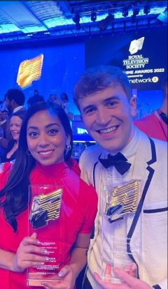 RWCMD grads Anjana, in a red dress, and Calum, in a wuite tuxedo, hold their Baftas while sming at the camera in a selfie style photo 
