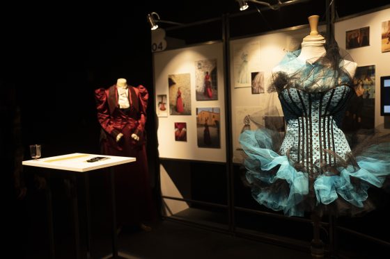 From the Balance exhibition, a manequin in the foreground in a pale blue corset and tulle skirt, with another manequin in a crimson victorian dress in the background. To the side are photos of the artists work 