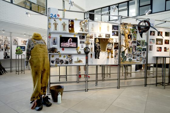 The Southern foyer at RWCD with a manequin with a pari of dirty yellow dungarees on, with an exhibition stand full of images and models showing the artists work 