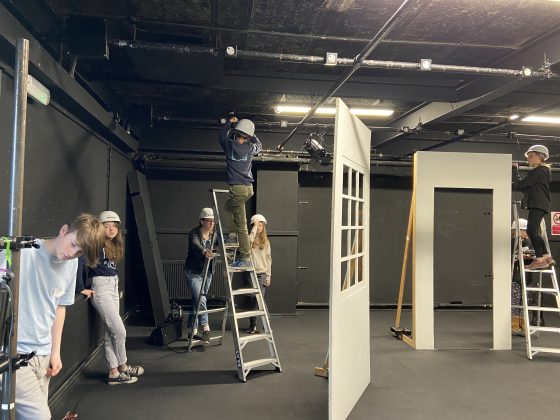 students working on the set, with one up a step ladder in a hard hat with two other students holding it steady. Other pieces of set - a window and a door - in the foreground