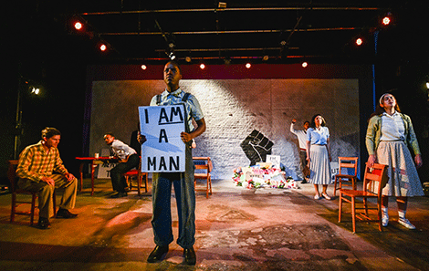 A student dressed in 1940’s clothes stands at the front of the stage with a placard saying ‘I am a Man’, watched by his fellow cast members  