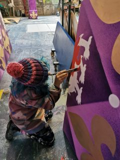 Millie, in a bobble hat, is painting a pantomime set in purple and gold 