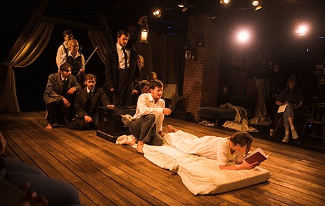 Millie designed both set and costume for RWCMD’s production of 'Indecent,' A girl lies in a white nightie on a bed reading a book, with a group of men and women behind her 